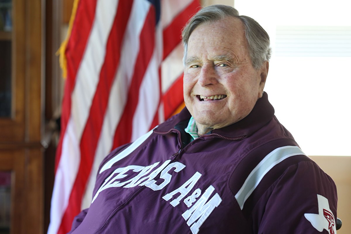 President George HW Bush seated wearing a Texas A&M jacket in front of an American flag