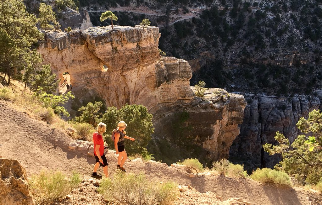 Two people wearing summer clothes and walking down and on paved trail in the background I sculpted limestone ridge with several natural windows.