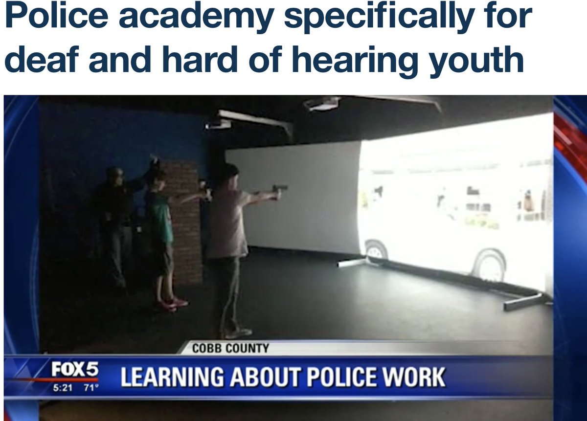 White presenting Deaf/Disabled children pointing guns at a projector screen with a car, people, building, and other visuals while a police officer looks on while leaning against a wall in a dark room.
