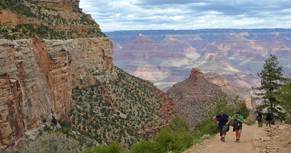 Image description: two hikers climbing up a steep, unpaved trail with a sheer cliff on their left, and several buttes and peaks in the distance.