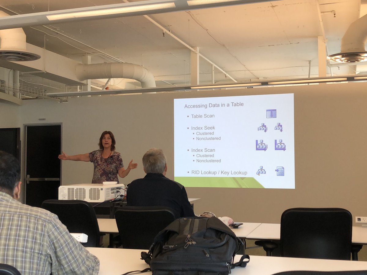 a white woman with brown hair gesturing with arms wide in front of an audience. the slide behind her is titled “Accessing Data in a Table” and contains bullet points about table scans, index seeks, index scans, and RID lookups/key lookups.