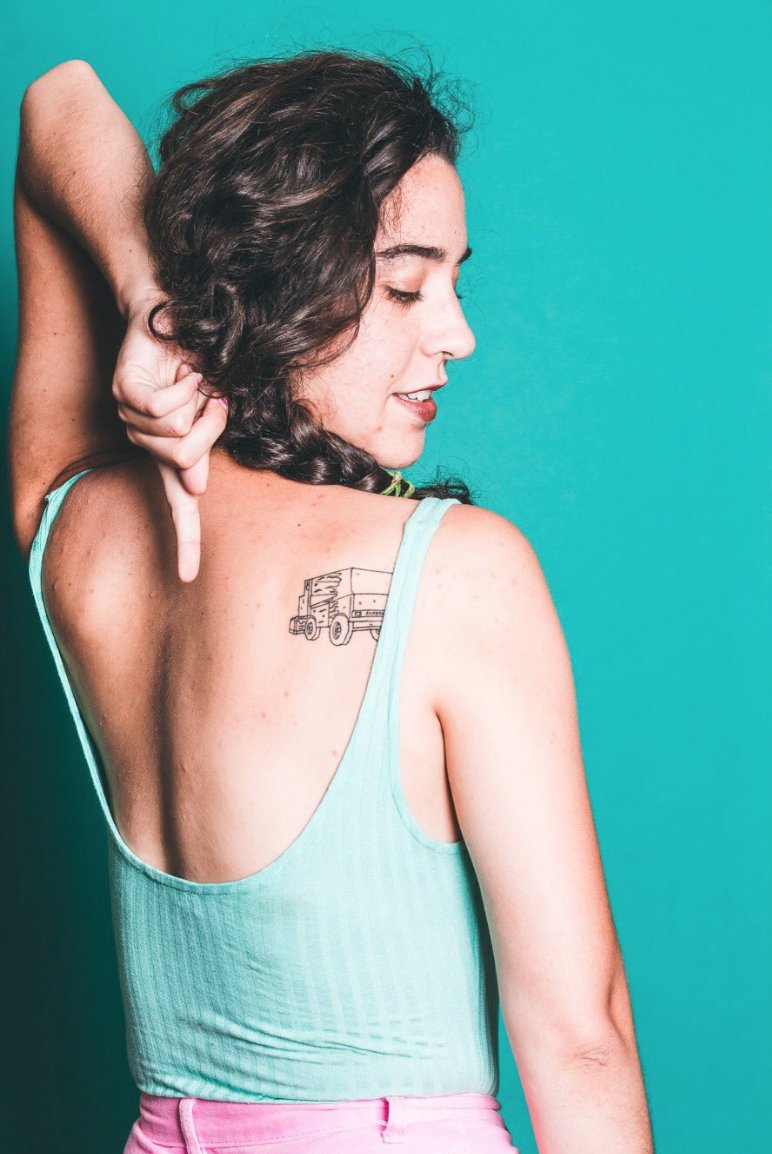 A white woman in a tank top with her back toward the camera, pointing her left arm at a tattoo of a Zamboni ice resurfacer near her right shoulder.