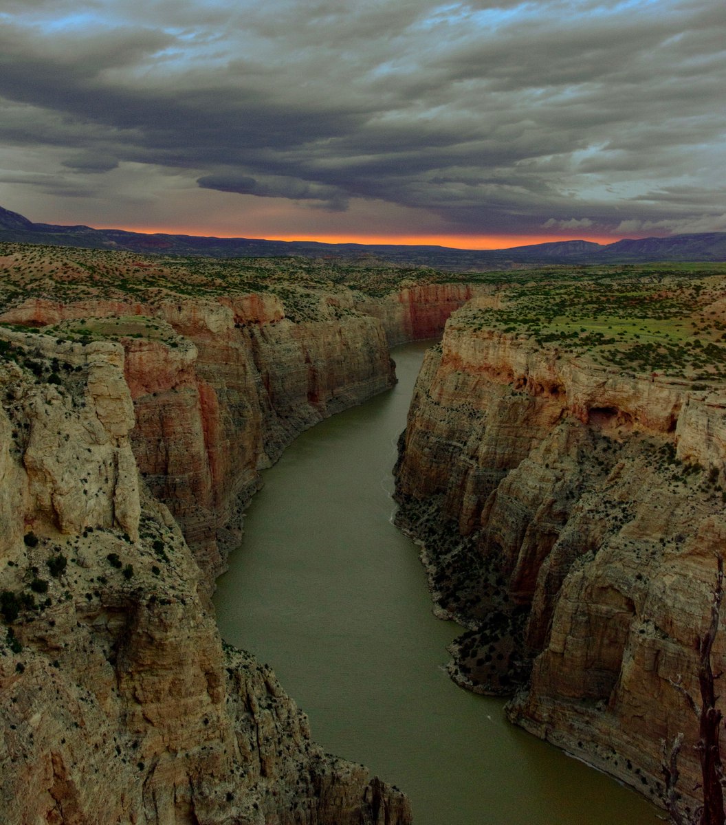 A river flows between the high walls of a narrow rock canyon under a stormy sunset sky.