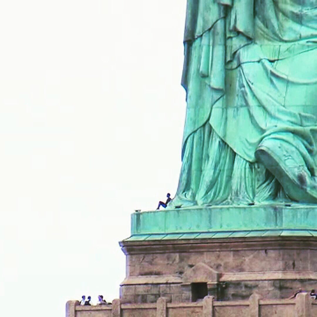 The base of the Statue of Liberty. A woman sits at the statue’s feet, with only her profile visible. Several people are below on the the ground.