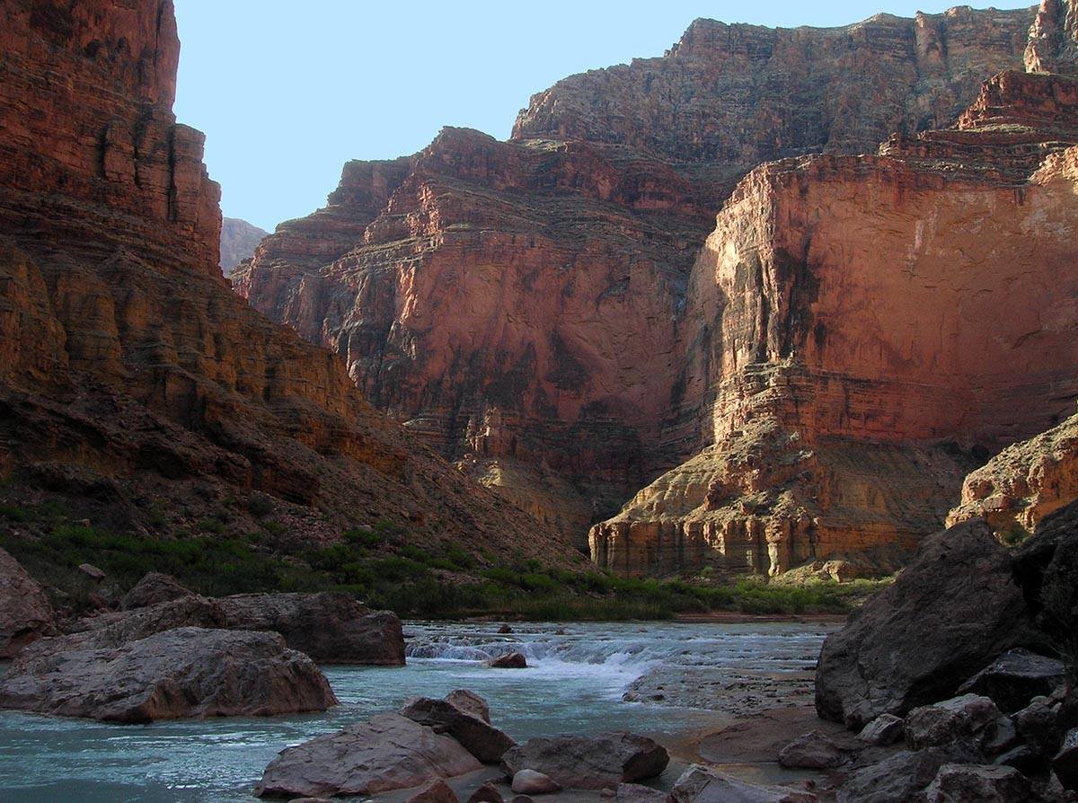 The blue water of the Little Colorado flowing over a gentle rapid, with the sun shining brightly on the towering cliffs beyond