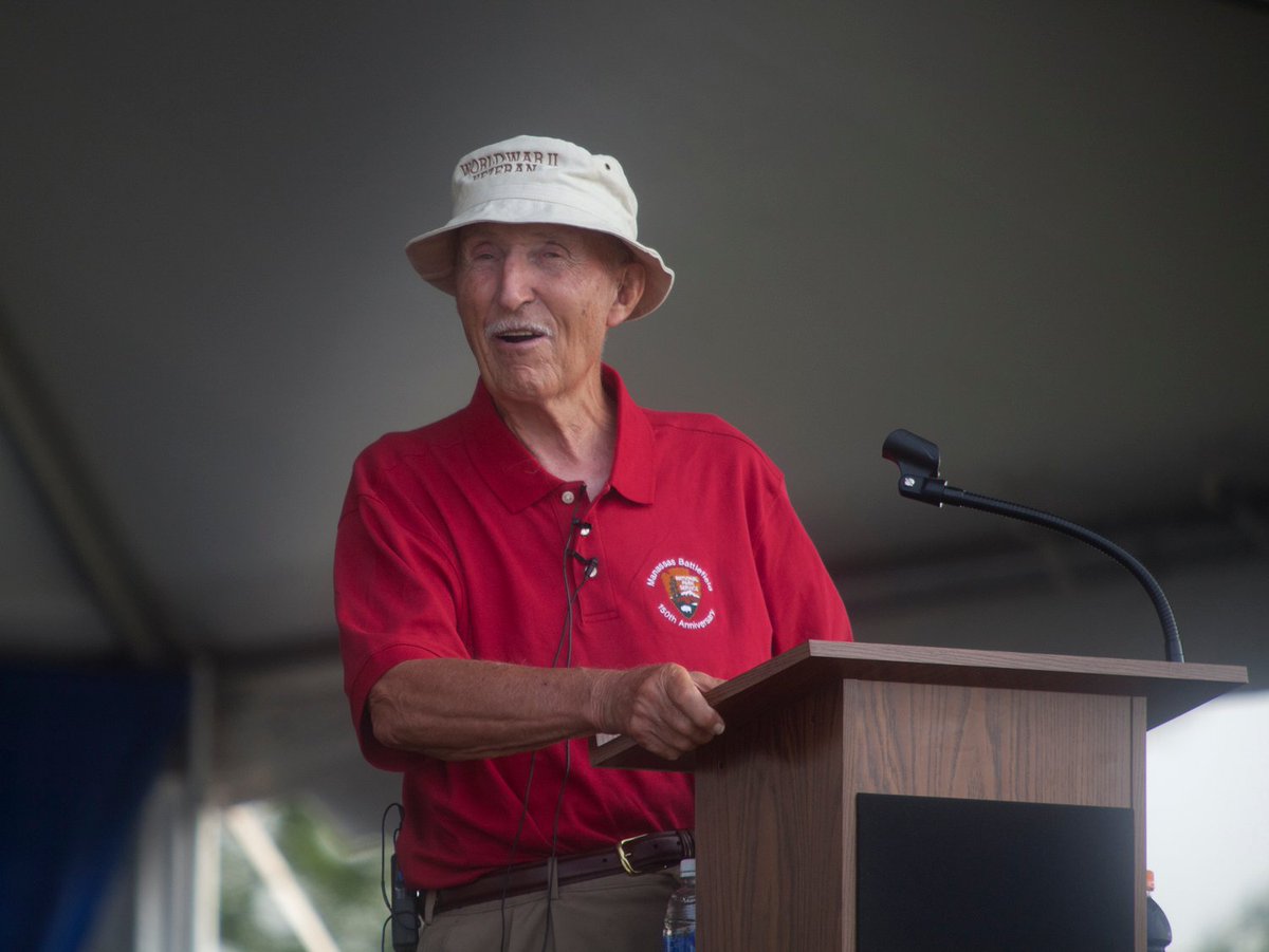 An older white man with a gray mustache wearing a floppy white hat and a red polo shirt speaks from behind a podium under a large tent.