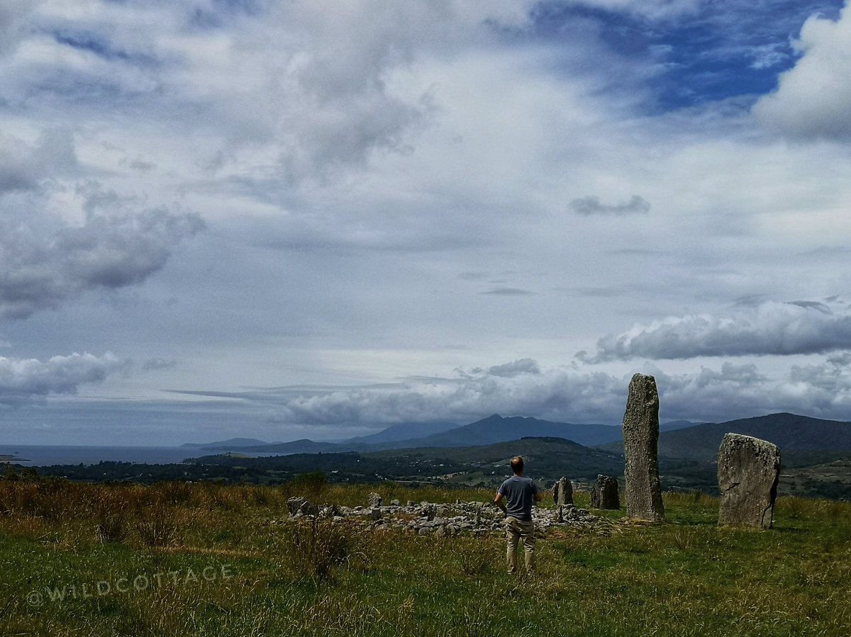 man standing outside of 2 very tall standing stones, smaller stone circle in front. mountains and sea in distance