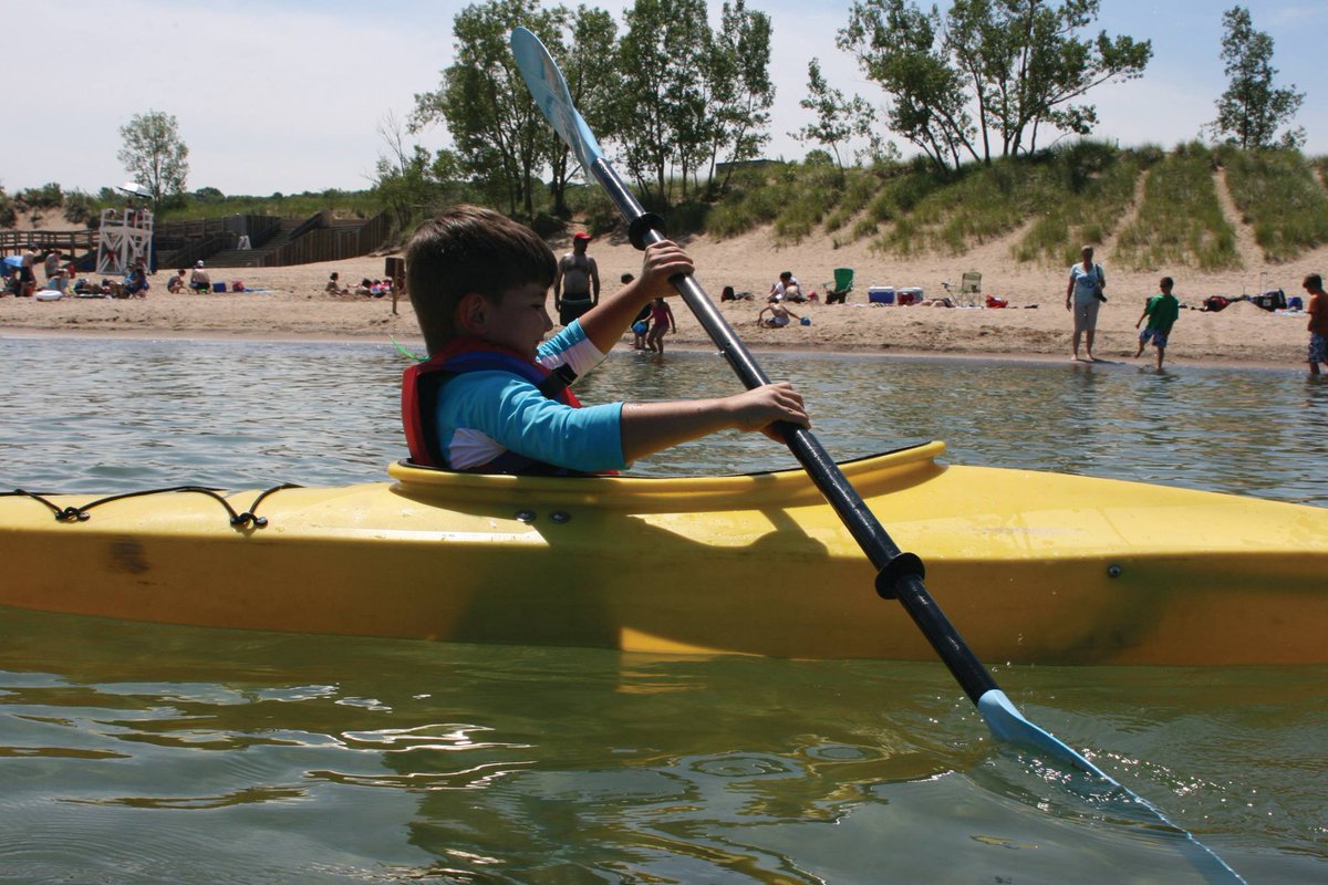 A young white boy with brown hair paddles a yellow kayak on a lake with people on the beach nearby watching him.
