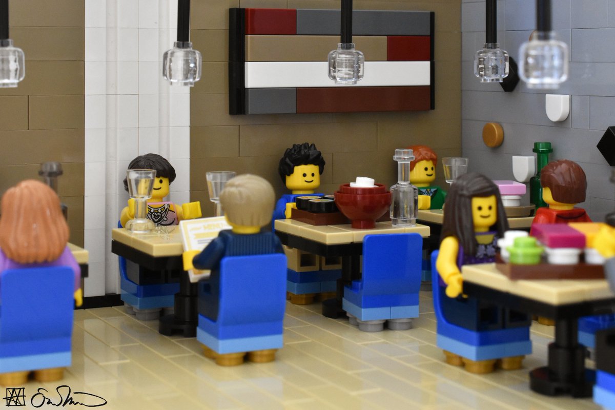 LGS sits at a restaurant table by himself and is surrounded by tables filled with couples. His table is filled with lots of food.