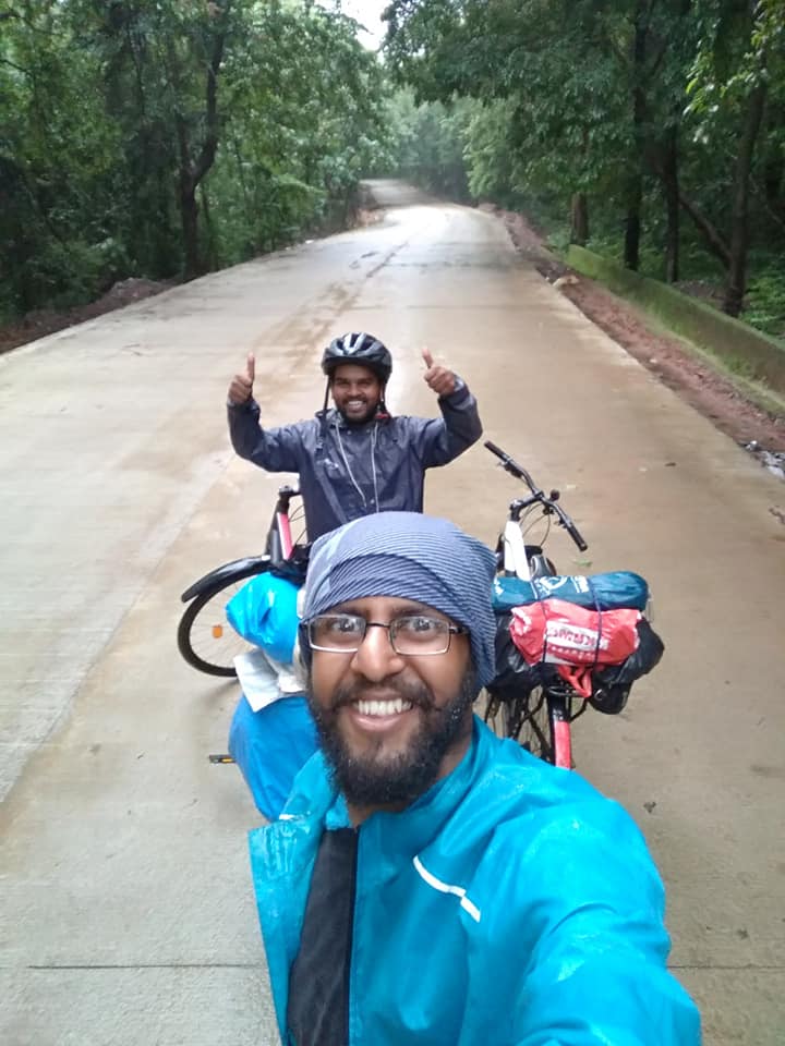 A boy taking a selfie with two bikes in the background. Another boy is standing behind him and giving two thumbs up.