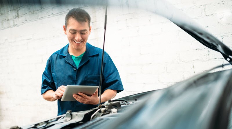 man smiling at tablet while standing in front of a car which has an open hood