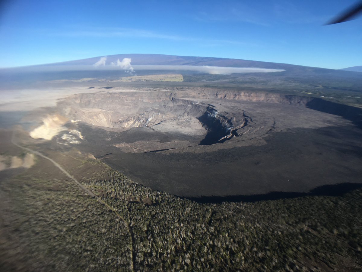 In this overflight image, HVO sits on the bluffs on the far side of the crater in the center of the view. Mauna Loa and the still-burning brushfire on its slope can be seen in the background.