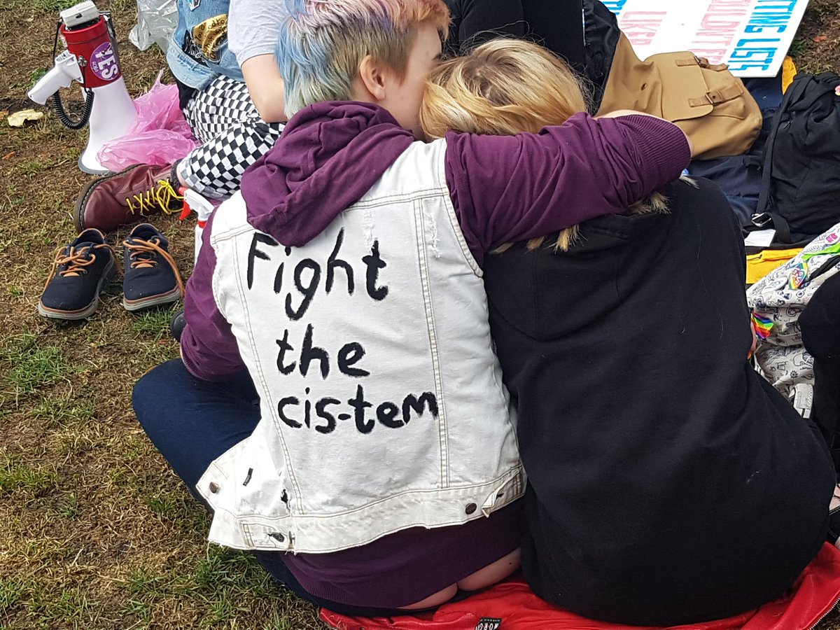 Two friends hug, one wearing a jacket that says 'Fight the cis-tem' on the back