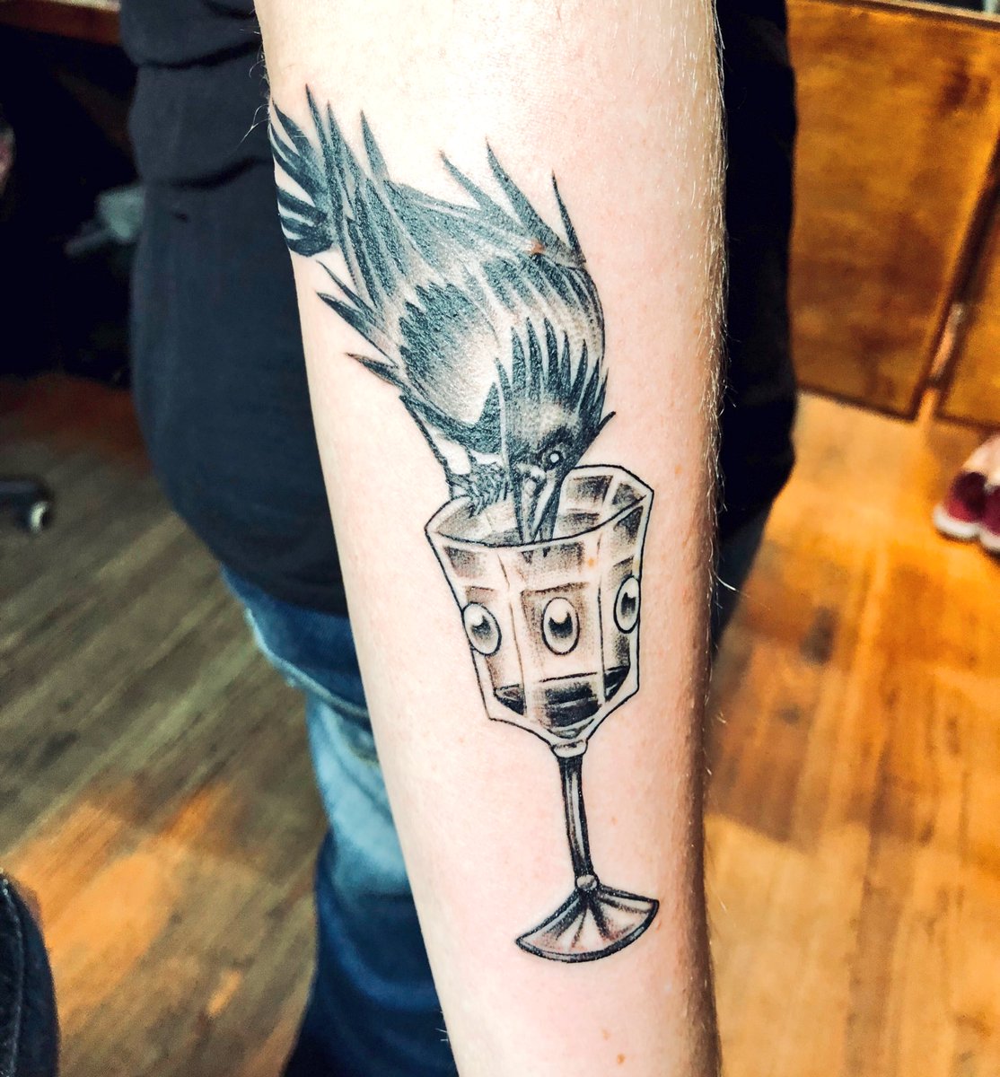 A photo of my right forearm, bearing a fresh tattoo. My skin is fair and the tattoo, which is done in black and gray, depicts a spiky-feathered crow perching on the rim of a bejeweled glass goblet. The crow is leaning in as though reaching to drink the small amount of liquid in the glass.
