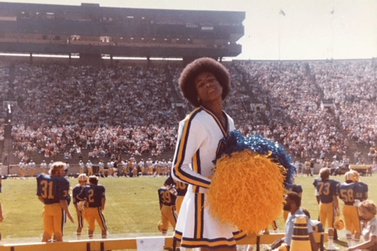 Cynthia Marshal at Berkeley in a cheerleading outfit with pompoms and the Cal football team on the field and fans in the stands.