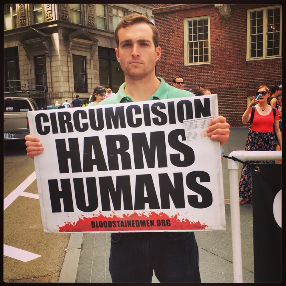 A.young man in his twenties holding a protest sign in a historic district of Boston that reads "Circumcision Harms Humans". Numerous observers are in the background, one is taking a picture with her cell phone. Bloodstainedmen.org