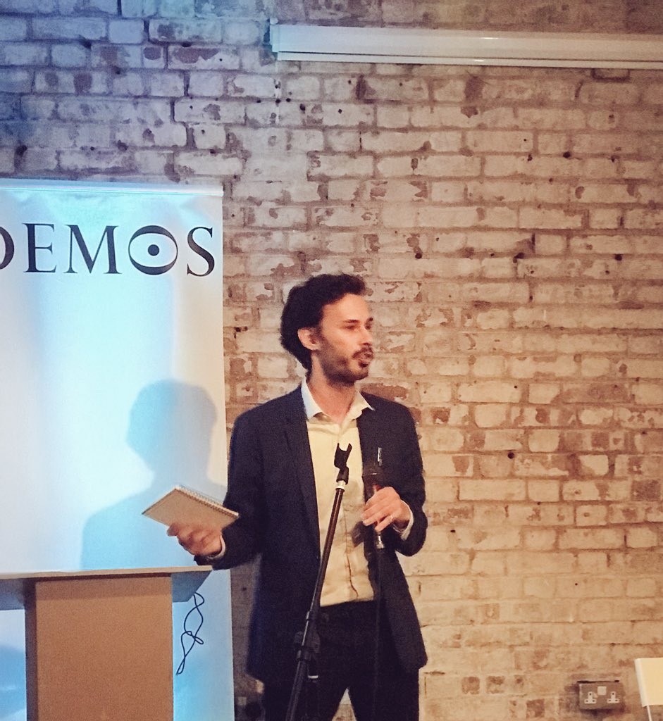 Carl Miller holding notepad in front of Demos sign addressing room.