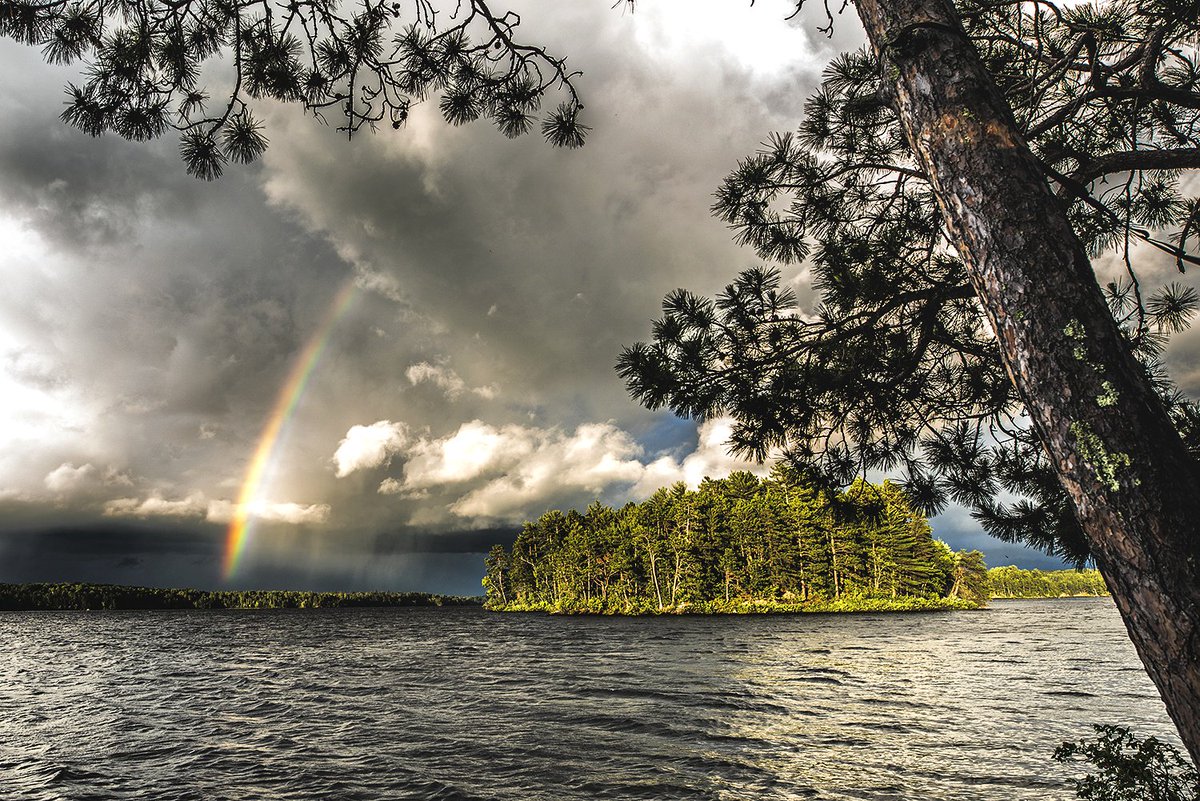 A view across a large lake shows a small island covered in thick green trees and a rainbow and a storm in the sky.