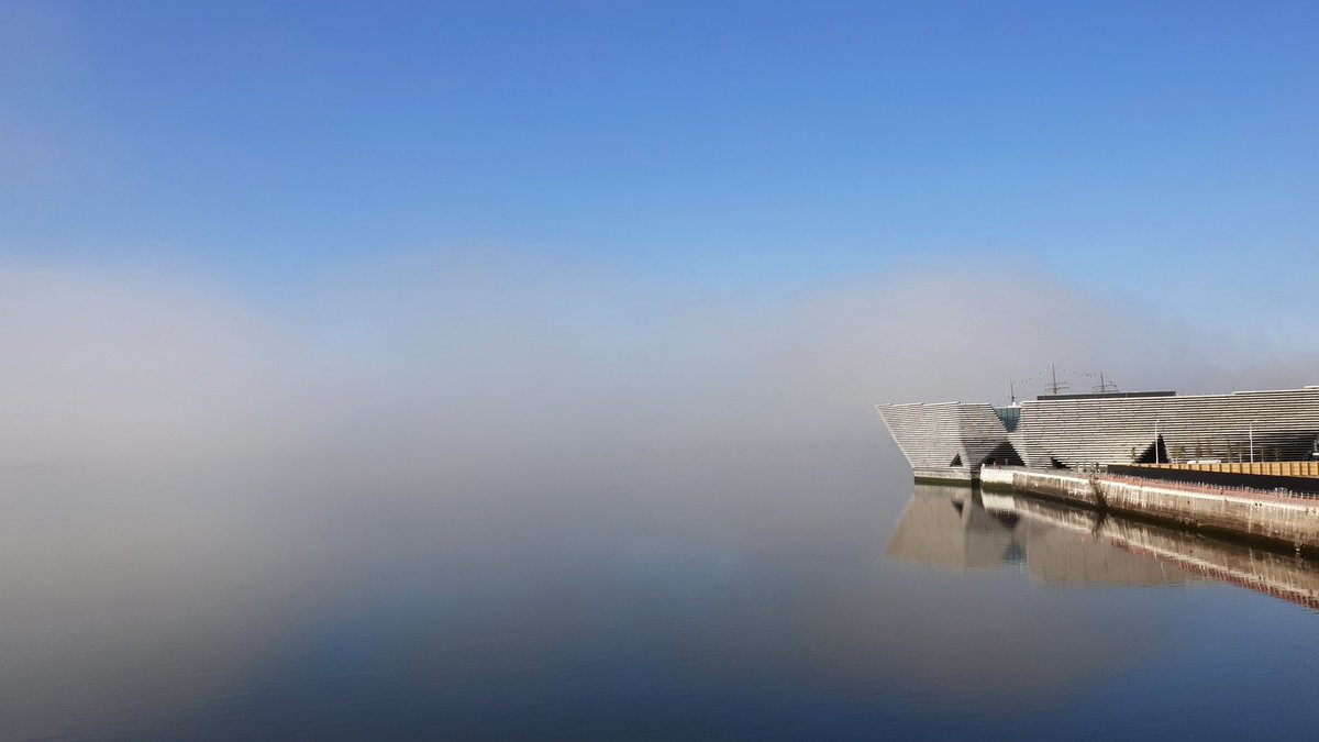 V&A Dundee on the water's edge. mist is in and obscures the horizon and land beyond so it looks like the museum is flowing in thin air. blue water and sky fringe the top and bottom edges of the misty image.