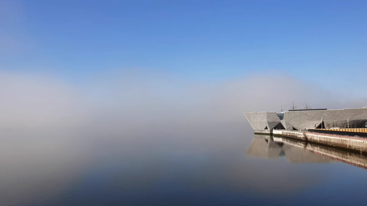 V&A Dundee on the water's edge. mist is in and obscures the horizon and land beyond so it looks like the museum is flowing in thin air. blue water and sky fringe the top and bottom edges of the misty image.