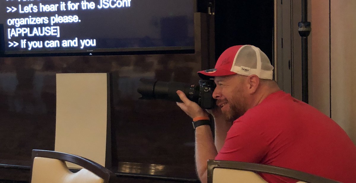 Derek Lindahl, organizer of JSConf, taking a photograph of the stage from a position in front of the live captioning screen which says, "let's hear it for the organizers"