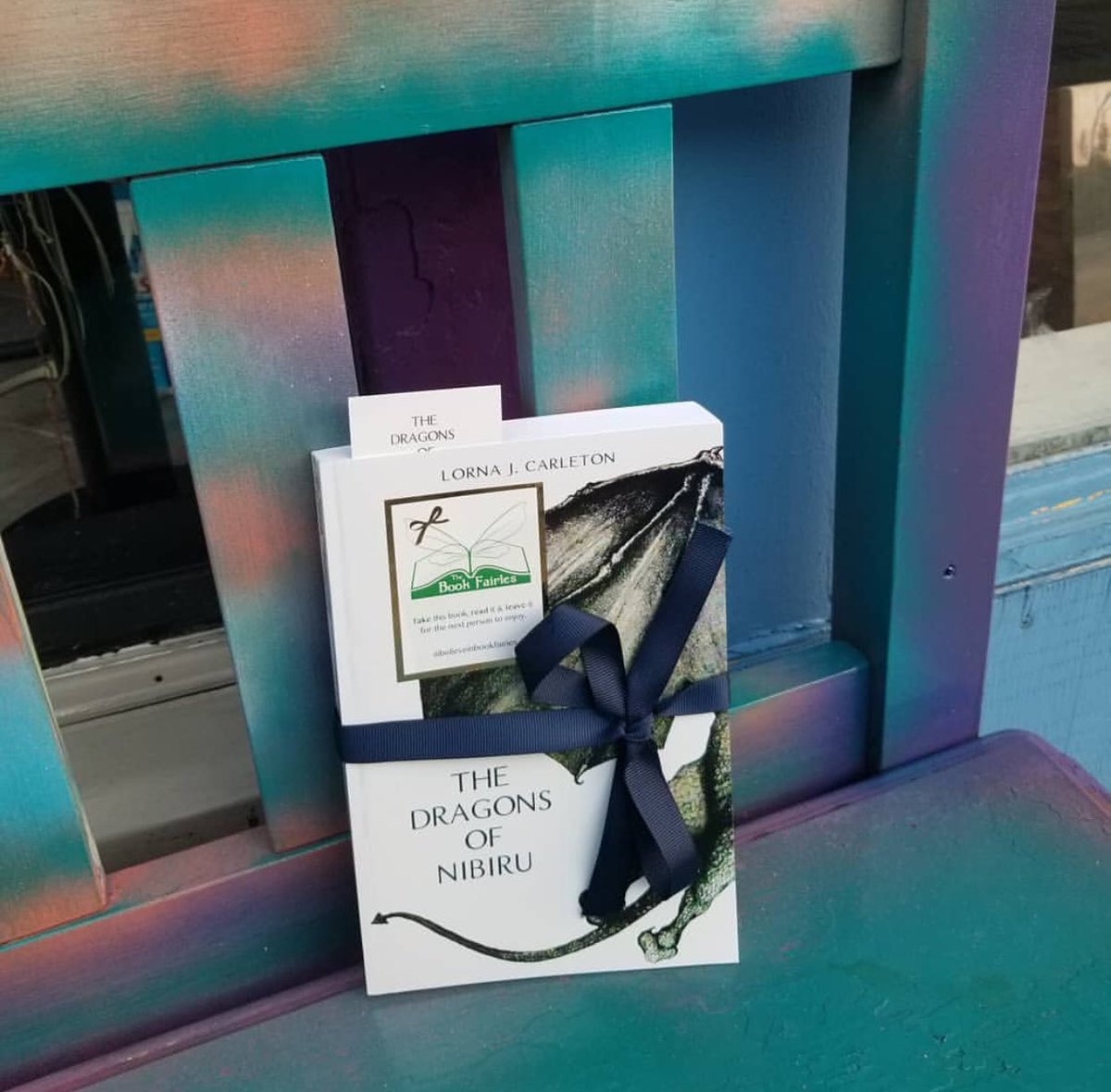 BookFairies_TO's tweet photo. This signed copies of Canadian author, Lorna J. Carleton’s The Dragons of Nibiru 🐉 is waiting on this beautifully coloured bench in downtown Milton for a lucky reader to find! 💚🐉
#ibelieveinbookfairies
#thedragonsofnibiru #dragonsofnibiru #fantasybooks https://t.co/KDzHvK1T5k