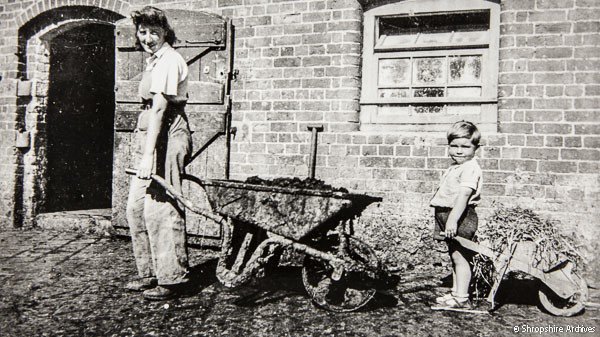 Here’s a Land Army Girl holding a wheelbarrow, being helped by a boy and his child-sized version, ca. 1943 at Alcaston Manor Farm, nr Church Stretton [PH/S/14/1/280] #Wheelbarrows