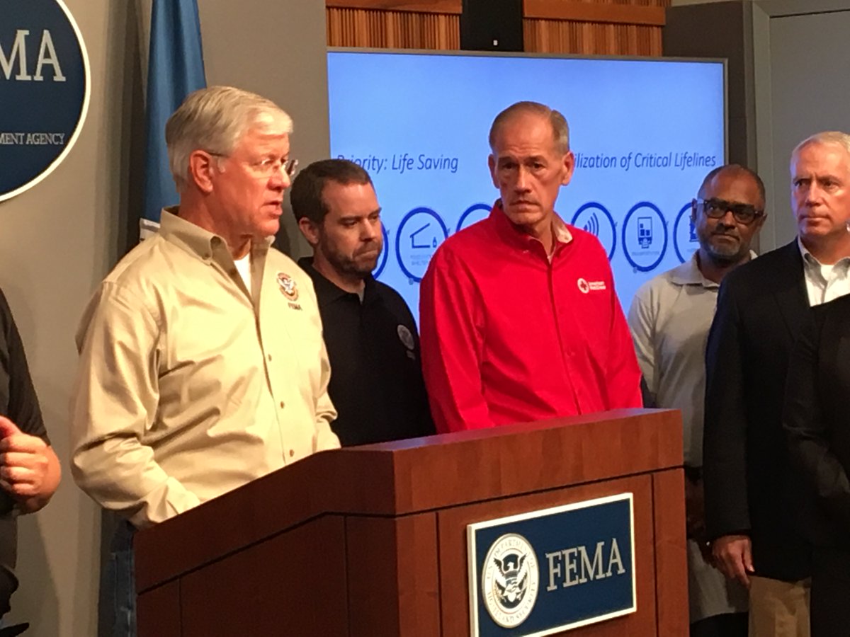 A man wearing a FEMA shirt stands at a press briefing podium that also has the FEMA seal on it. Next to him are four other men who are watching him as he speaks. In the background is a screen that shows several icons that are partially obscured, and the words "Priority: Life Saving" and "Stabilization of Critical Lifelines"