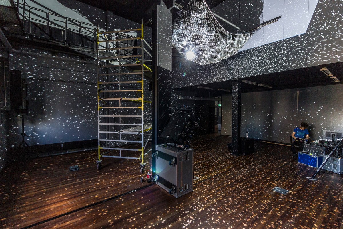 Boxy room with high ceilings and wooden floor covered in small reflections dotting all the surfaces. Some scaffolding in the room. A pile of disco balls in a net hanging from the ceiling.