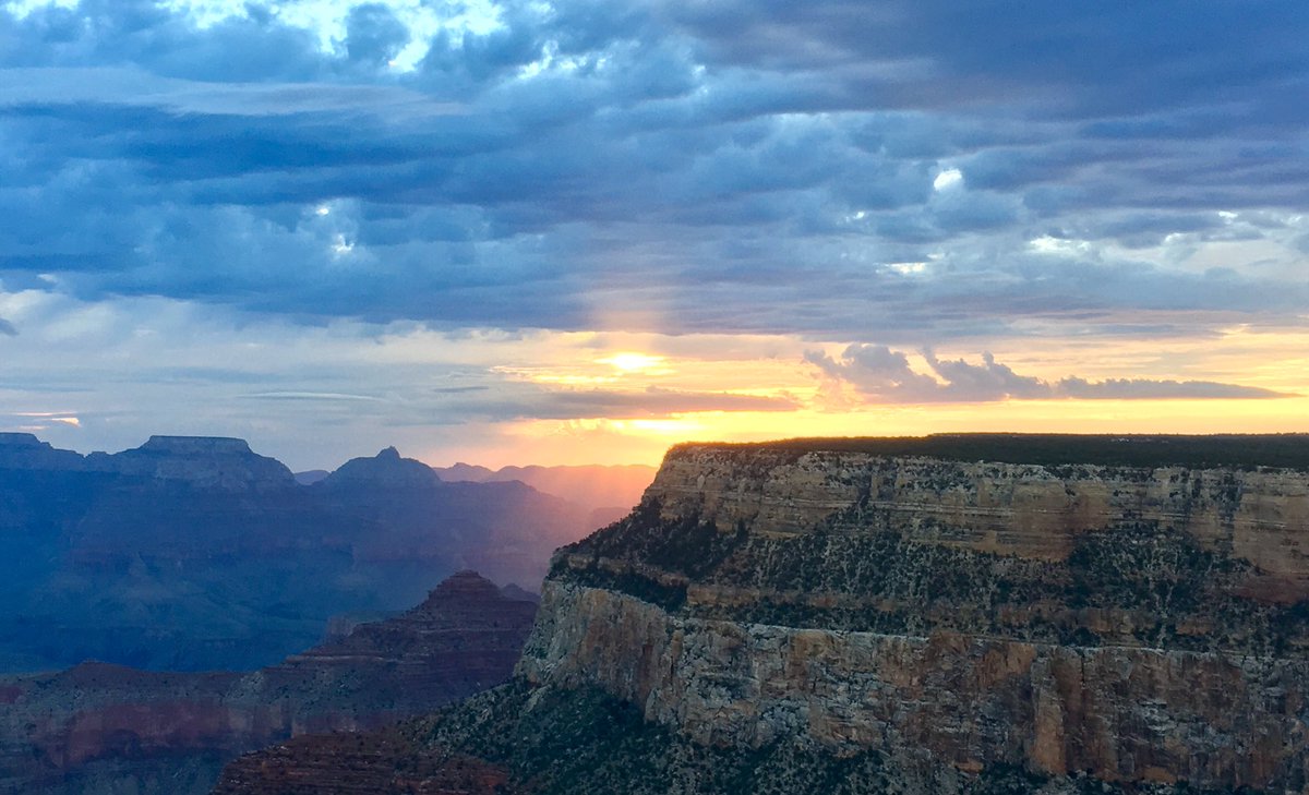 A massive cliff wall in the foreground, with yellow sunrise light filtering through layers of bluish clouds. Several peaks rising up from the canyon’s depths.