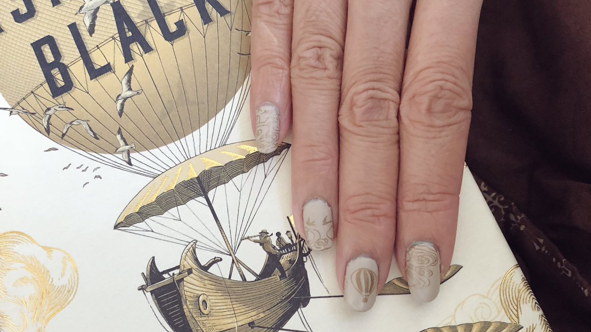 Closeup of cream and gold nail art with hot air balloon, cloud patterns and birds next to book cover of Washington Black