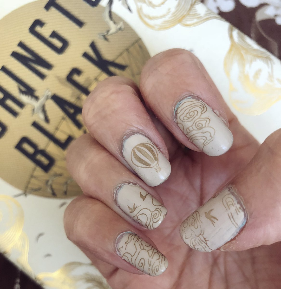 Closeup of cream and gold nail art with hot air balloon, cloud patterns and birds next to book cover of Washington Black
