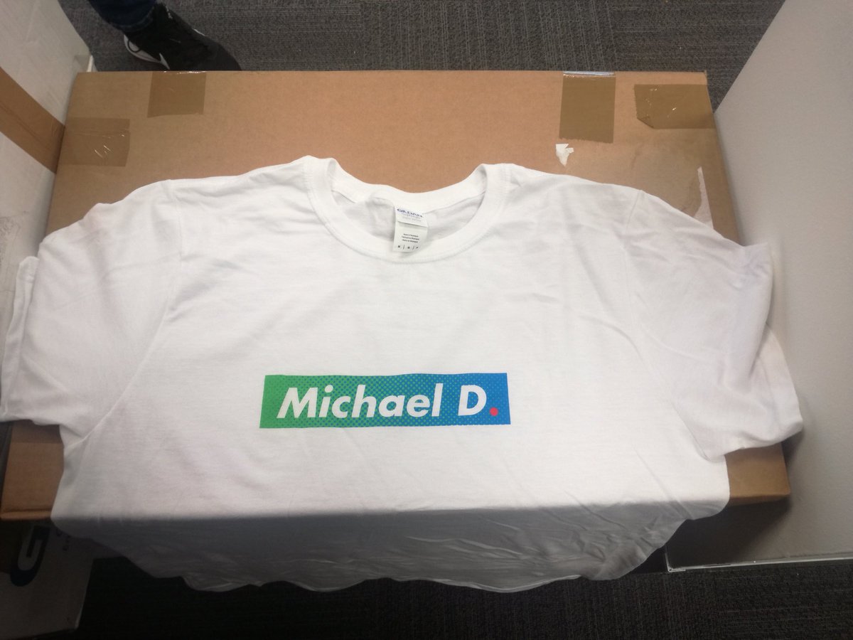A white t-shirt with the blue green campaign slogan that reads “Michael D” in white, with a red dot. The t-shirt is on an unopened cardboard box.