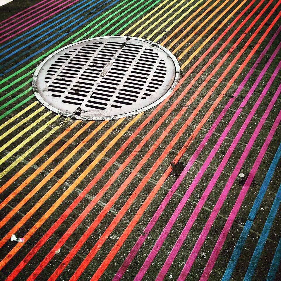 An image of a silver manhole cover on top of a rainbow colored pedestrian crosswalk.