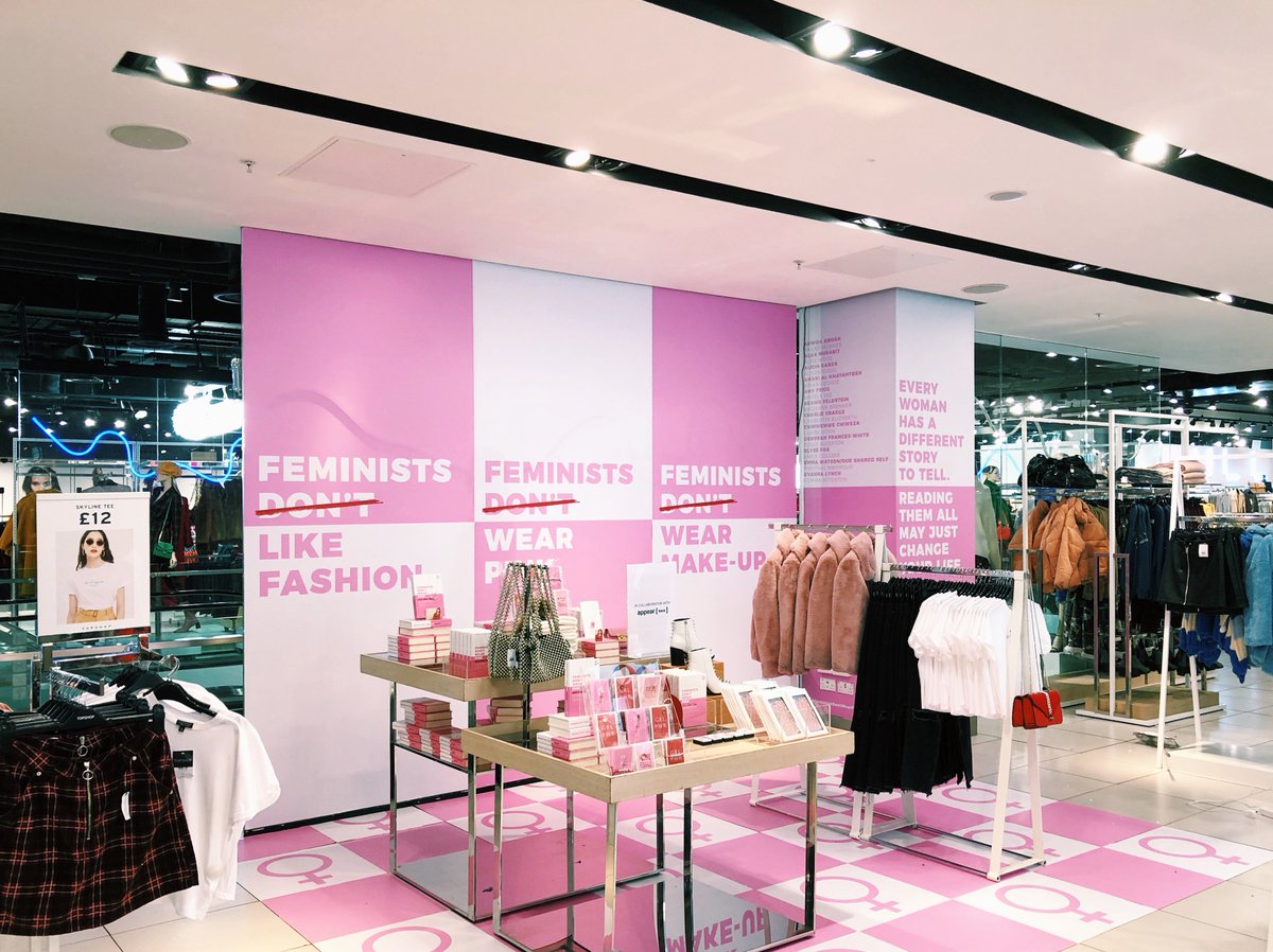 A photo of a pop-up stall within a Topshop store; there are several pink and white posters with bold slogans such as 'Feminists like fashion’, ‘Feminists wear make up’ and ‘Every woman has a different story to tell. Reading them all may just change your life’, alongside copies of the book, Feminists Don't Wear Pink.