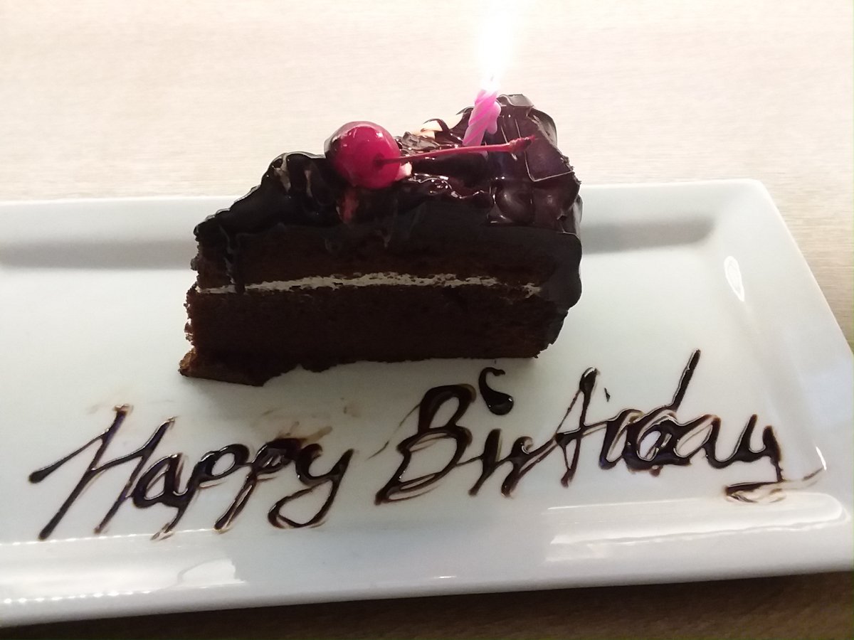 photo of a slice of dark chocolate layer cake with dark chocolate frosting with a layer of whipped cream in the center. It is topped with a red maraschino cherry and a small pink birthday candle which is lit. The cake sits on a rectangular white dish with “Happy Birthday” handwritten, in script, with chocolate icing.
