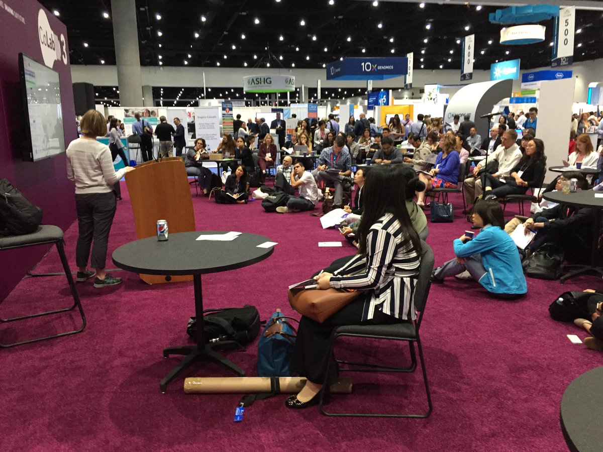 A person stands in front of a podium, looking at a TV screen with a presentation on it. The audience faces the screen, as well. They are sitting on the floor or in chairs at small circular tables.