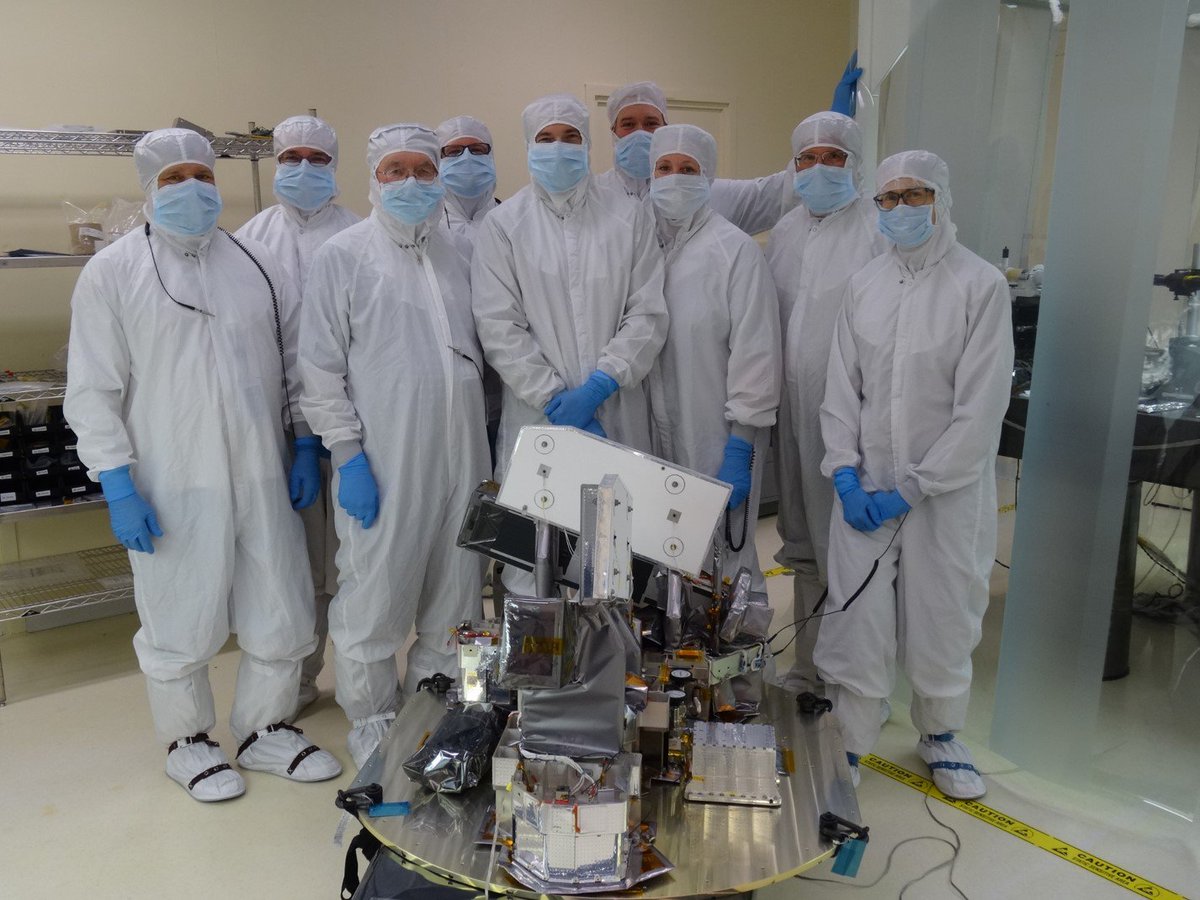 Members of the MIGHTI instrument team stand behind the instrument in a clean room, wearing bunny suits.