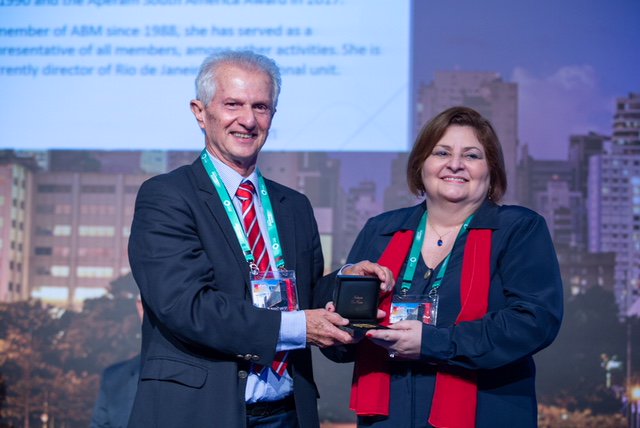 The picture shows a man and a woman holding a small black box. The woman wears a red scarf and has short hair.