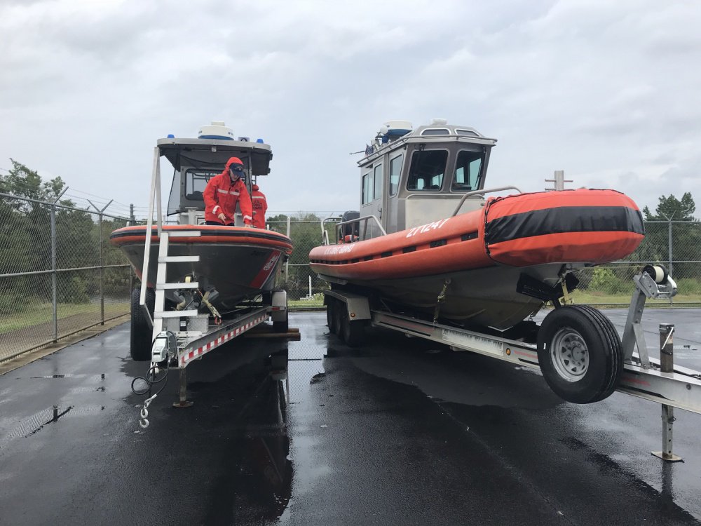 Two US Coast Guard boats are in a rainy parking lot, with two men working on them.