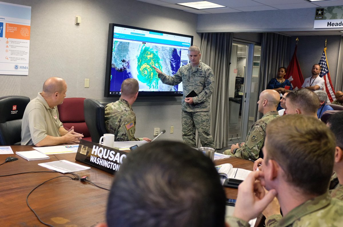 A conference room with US Army Corps of Engineer staffers participating in a meeting about Hurricane Michael. The storm is shown on a radar photo on a big screen and one of the staffers is pointing to it.