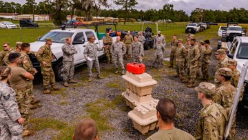 A large group of soldiers with the National Guard are positioned in a large circle in a gravel parking lot. In the middle is a stack of heavy duty equipment in cases.