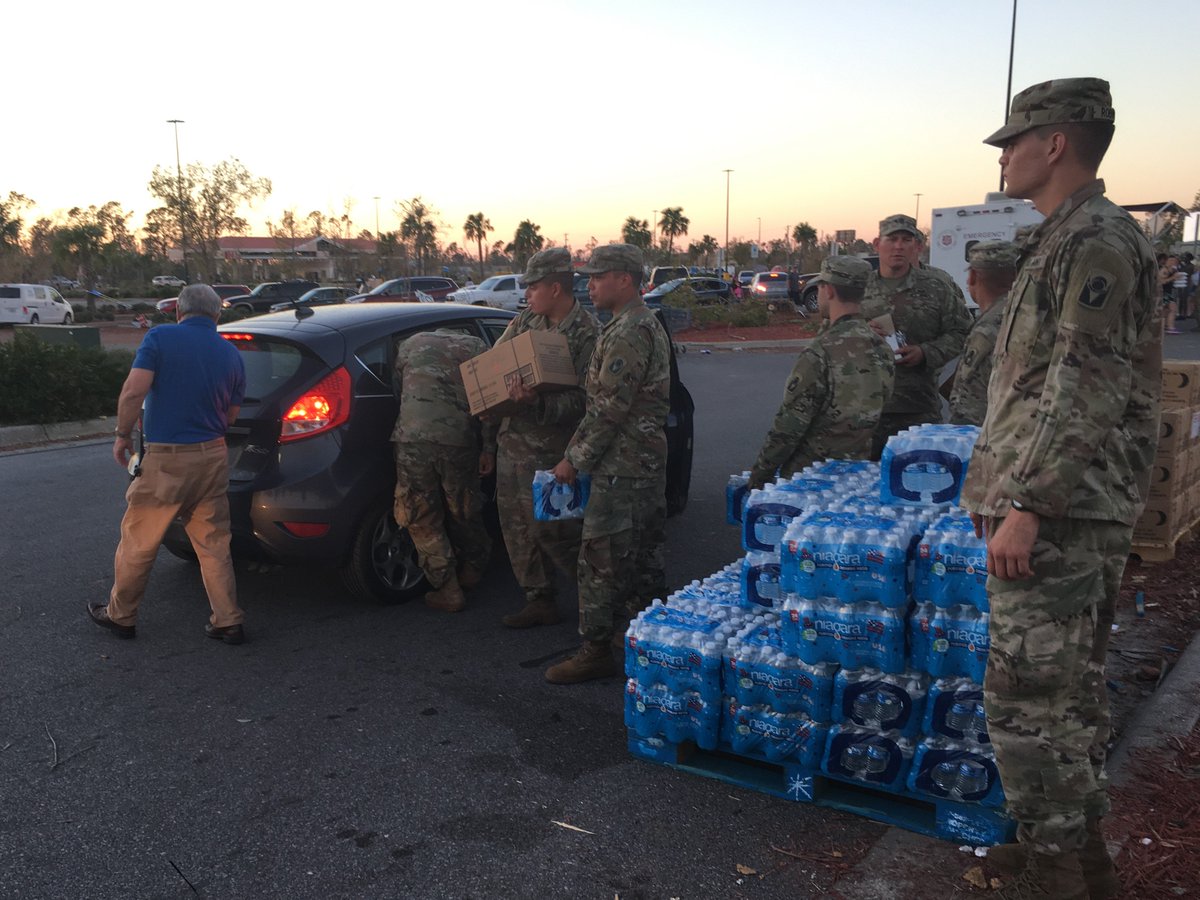 In a parking lot, seven soldiers wearing fatigues are standing by a large pallet of bottled water. A man wearing a blue polo and khakis is opening the trunk of his car, while three of the soldiers bring water and boxes of supplies to his car. A Salvation Army truck is in the background.