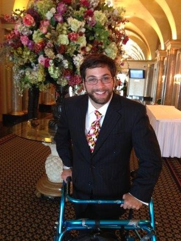 Image of Aaron Kaufman. He is standing in a large hallway with flowers on a table behind him. He is wearing a suit and using a walker.