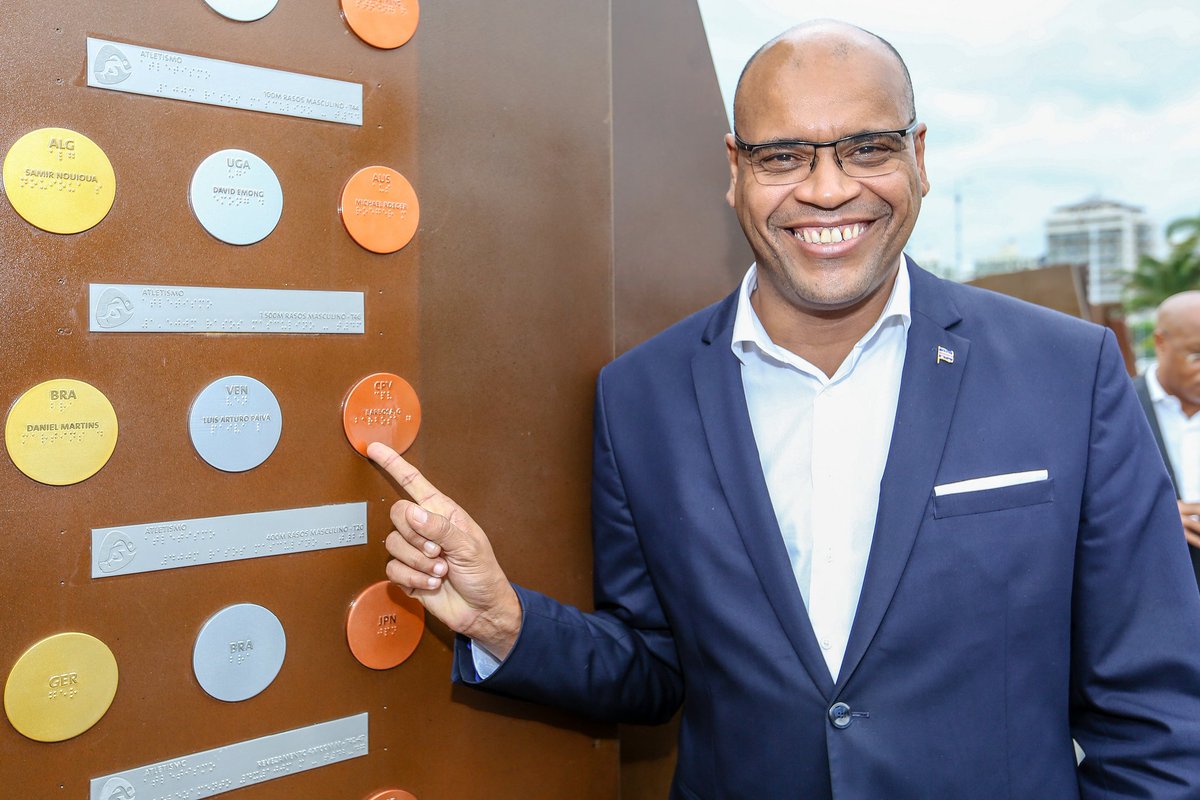 Minister Fernando Elísio smiles to the camera. He points to a board that has names of athletes who won medals.