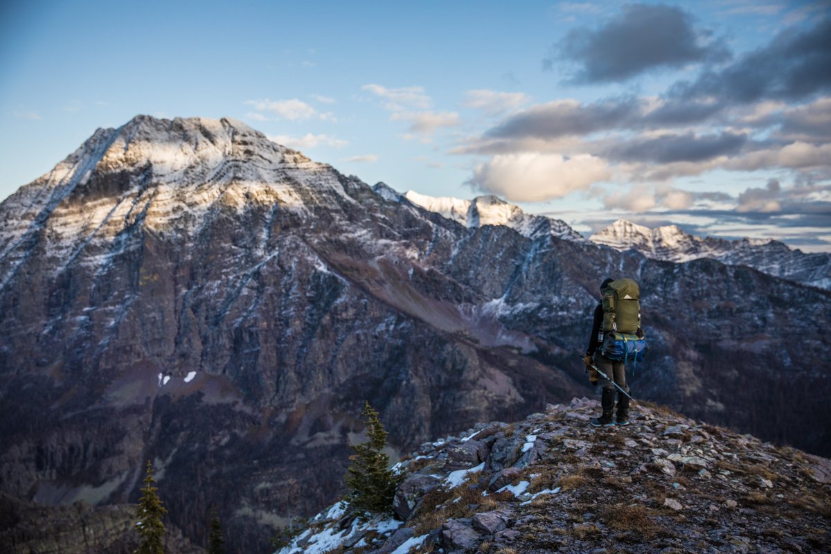 A backpacker stands on a hill with snowy rocky peaks in the background.