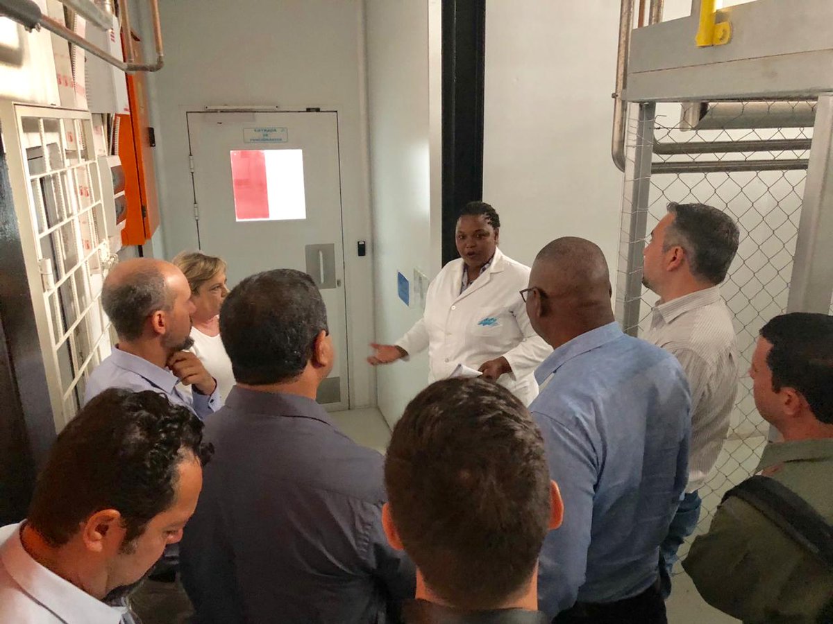 Several people are gathered in front of a lady wearing medical white coat. She talks in front of a door, as if showing the way.