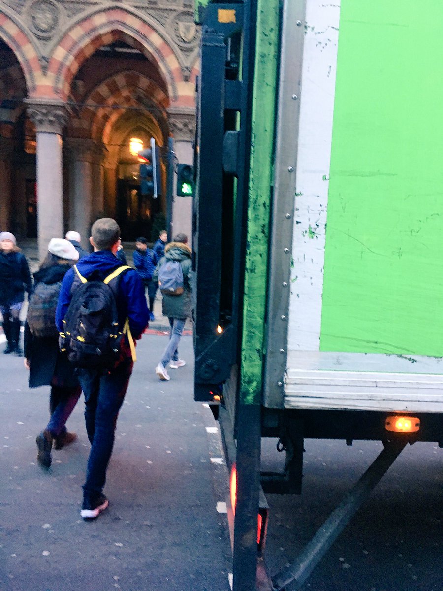 A large green lorry is blocking the pedestrian crossing, inhibiting Amit and Kika from crossing the road.