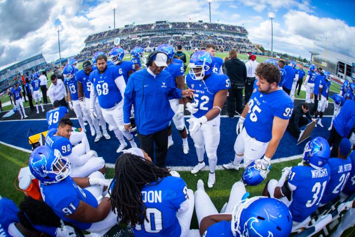 UB Football Team surrounding coach on football field.