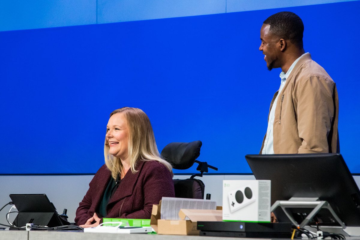 Jessica sitting at desk, with Jamal standing beside her - smiling in full flow demo mode. Laptops and Xbox in front of her and to the side